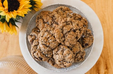 A plate of freshly baked cookies on a white plate. A vase of sunflowers is in the upper left corner.