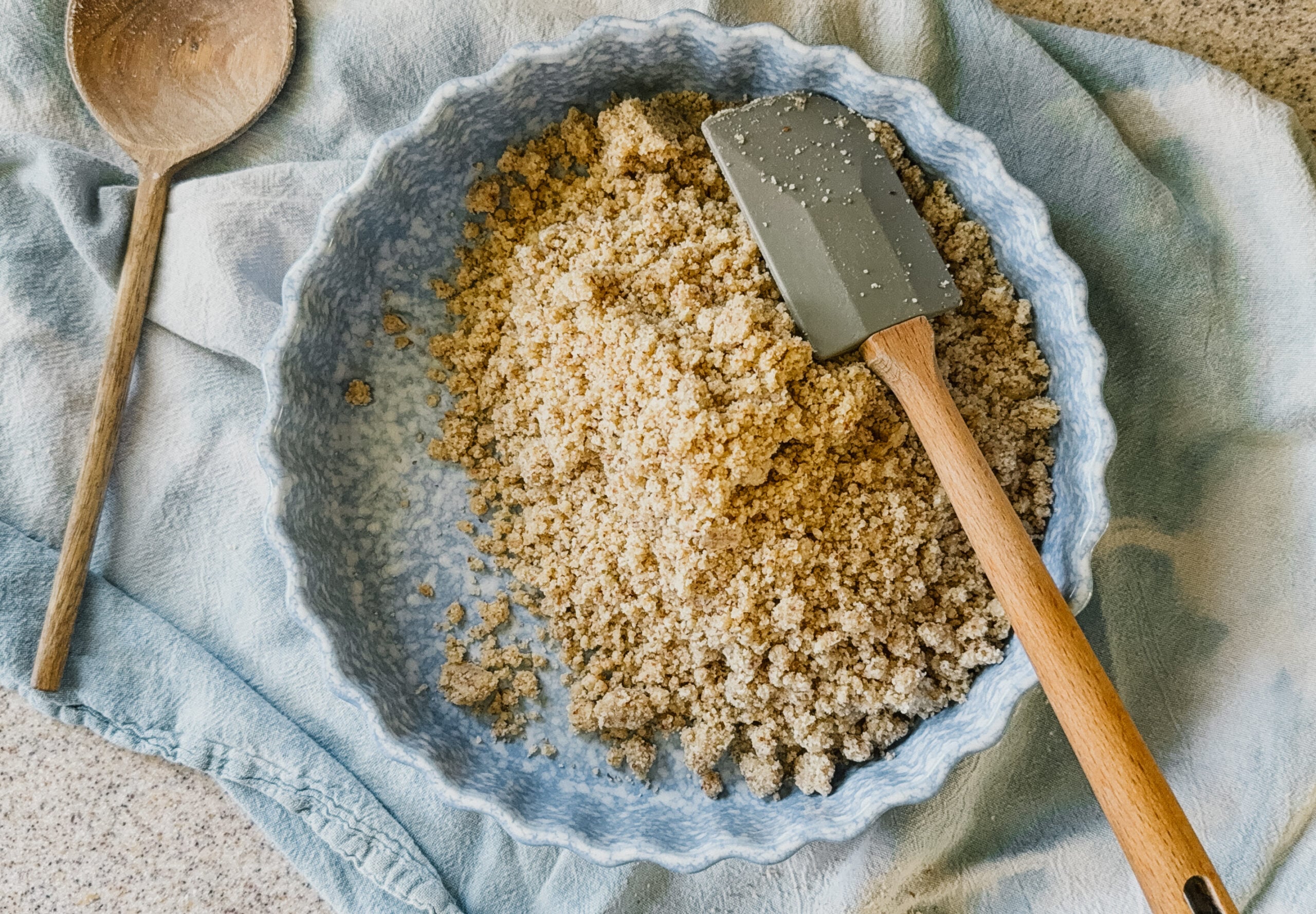 A blue pie plate filled with a pie crust mixture about to be pressed. The pie dish sits on top of a blue tea towel and a wooden spatula sits on its upper right side.