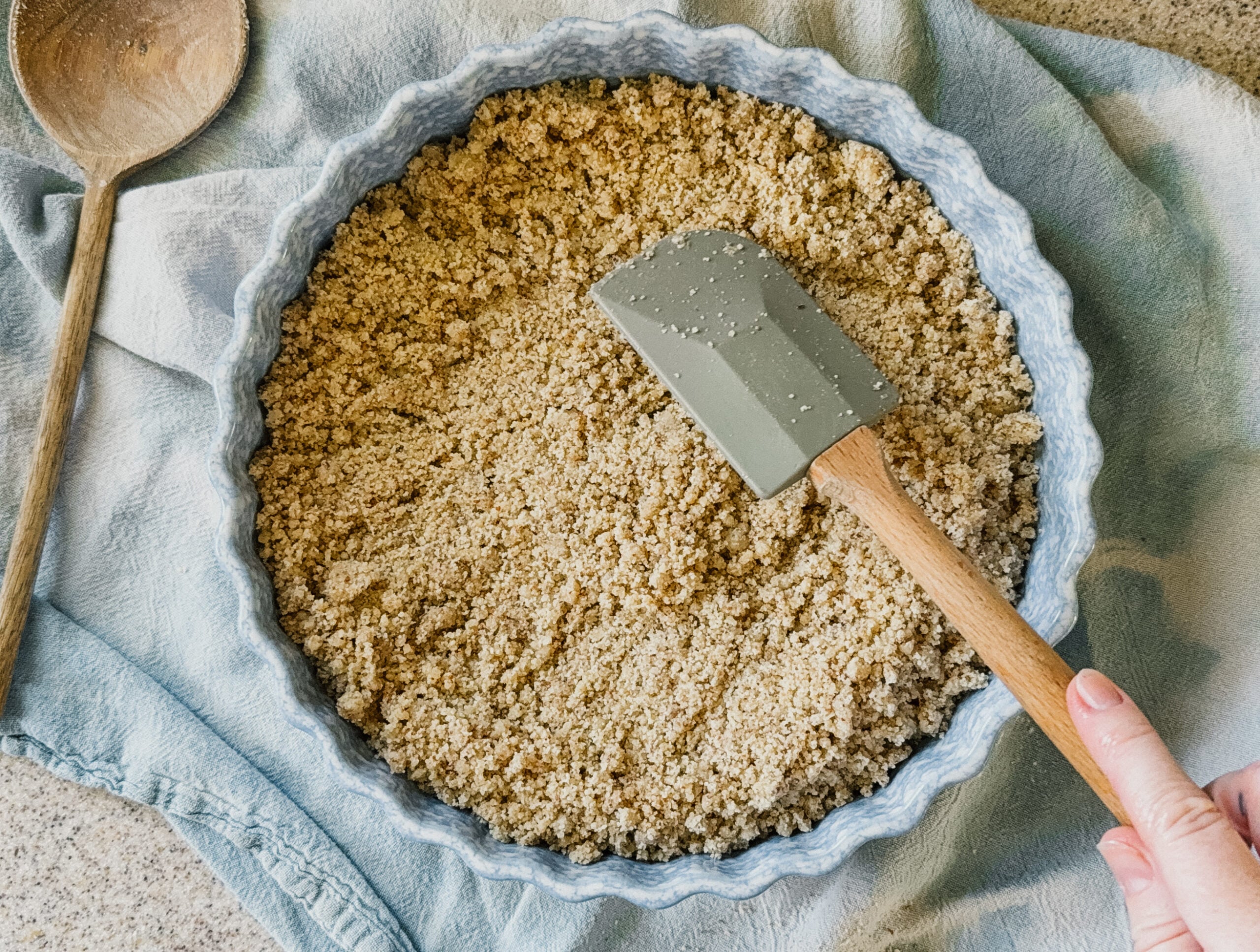 A spatula pressing a crust into a blue pie dish. The dish sits on top of a blue tea towel.