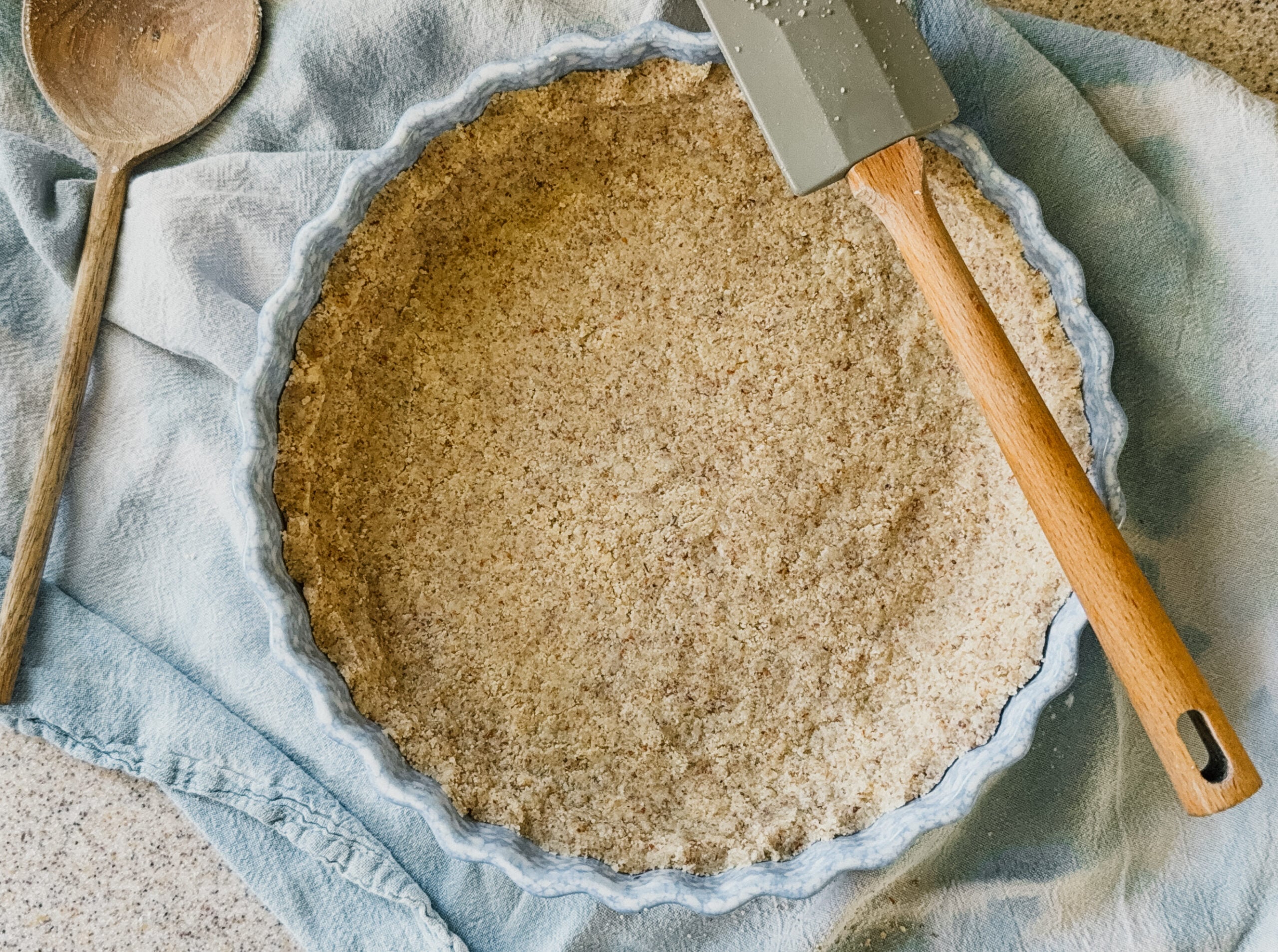 A blue pie plate filled with a pressed pie crust about to be baked. The pie dish sits on top of a blue tea towel and a wooden spatula sits on its upper right side.