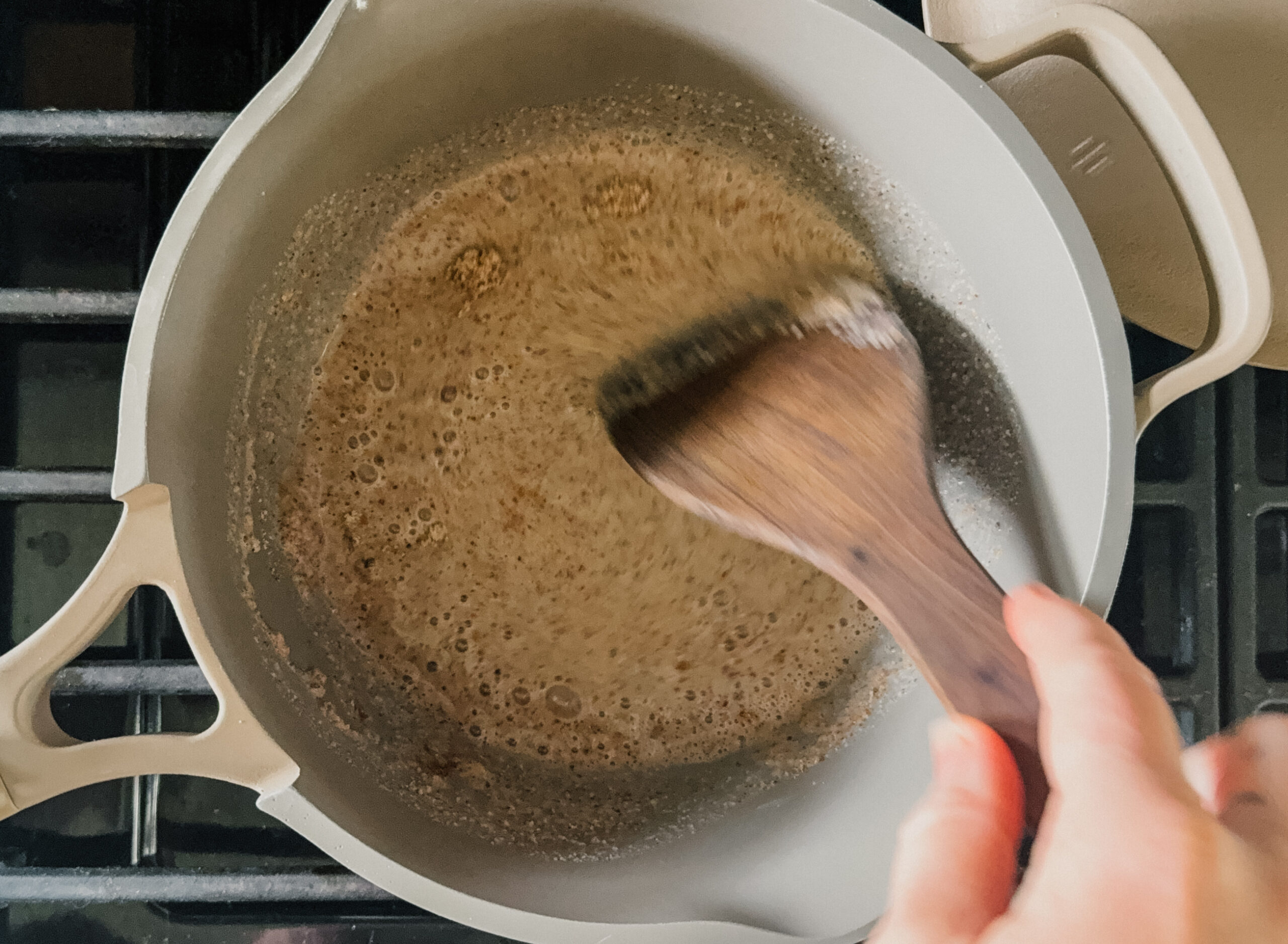 A hand holding a wooden spoon stirring a pot of porridge on a stove.