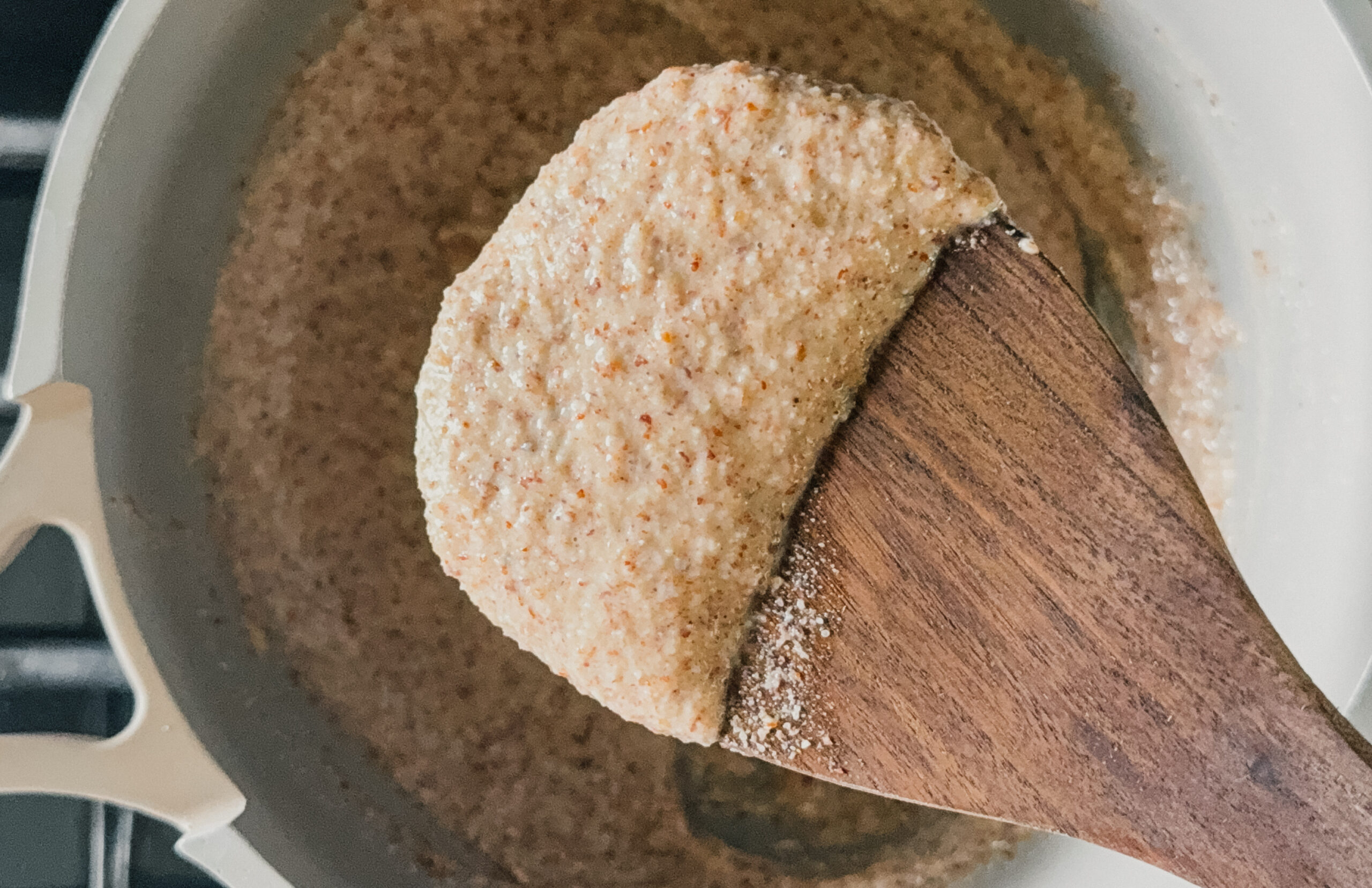 A wooden spoon with a scoop of porridge above a pot with the remaining porridge on a stovetop.