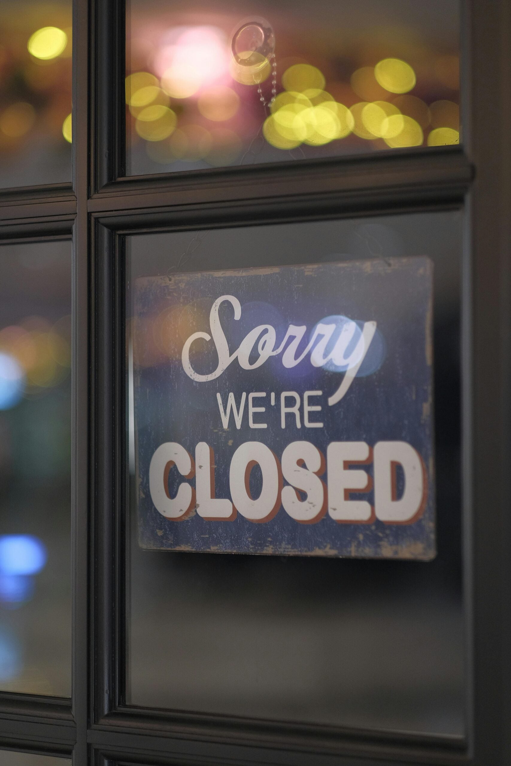 Close-up of a 'Sorry We're Closed' sign on a glass window with bokeh lights in the background.